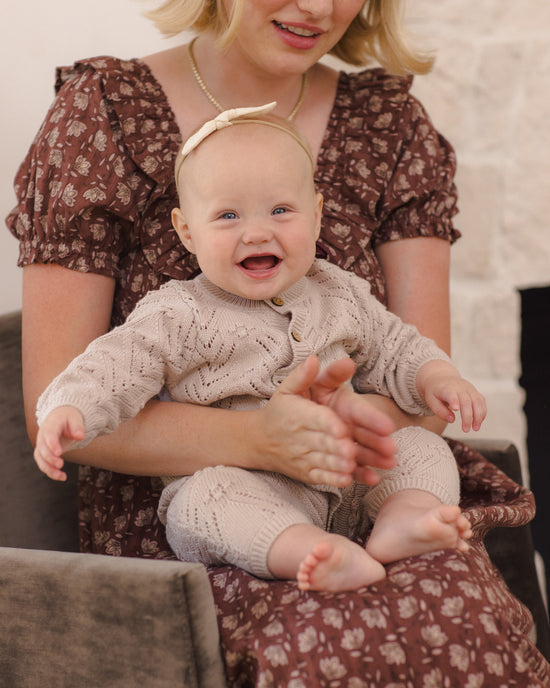 A baby wearing a beige cable-knit cardigan and matching knit pants sits on a woman's lap, smiling and clapping. The woman wears a brown floral dress.