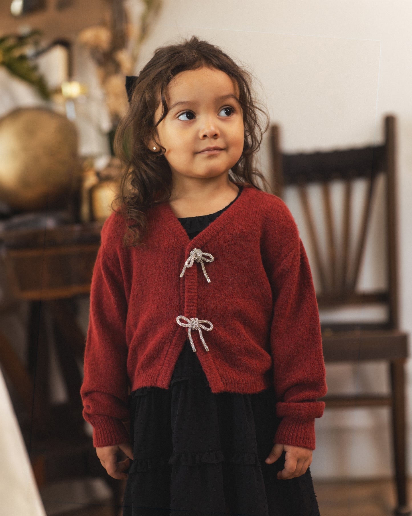 A young girl with long brown hair wearing a red knit cardigan with white tie closures over a black dress, standing in a softly blurred indoor room.