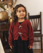 A young girl with long brown hair wearing a red knit cardigan with white tie closures over a black dress, standing in a softly blurred indoor room.