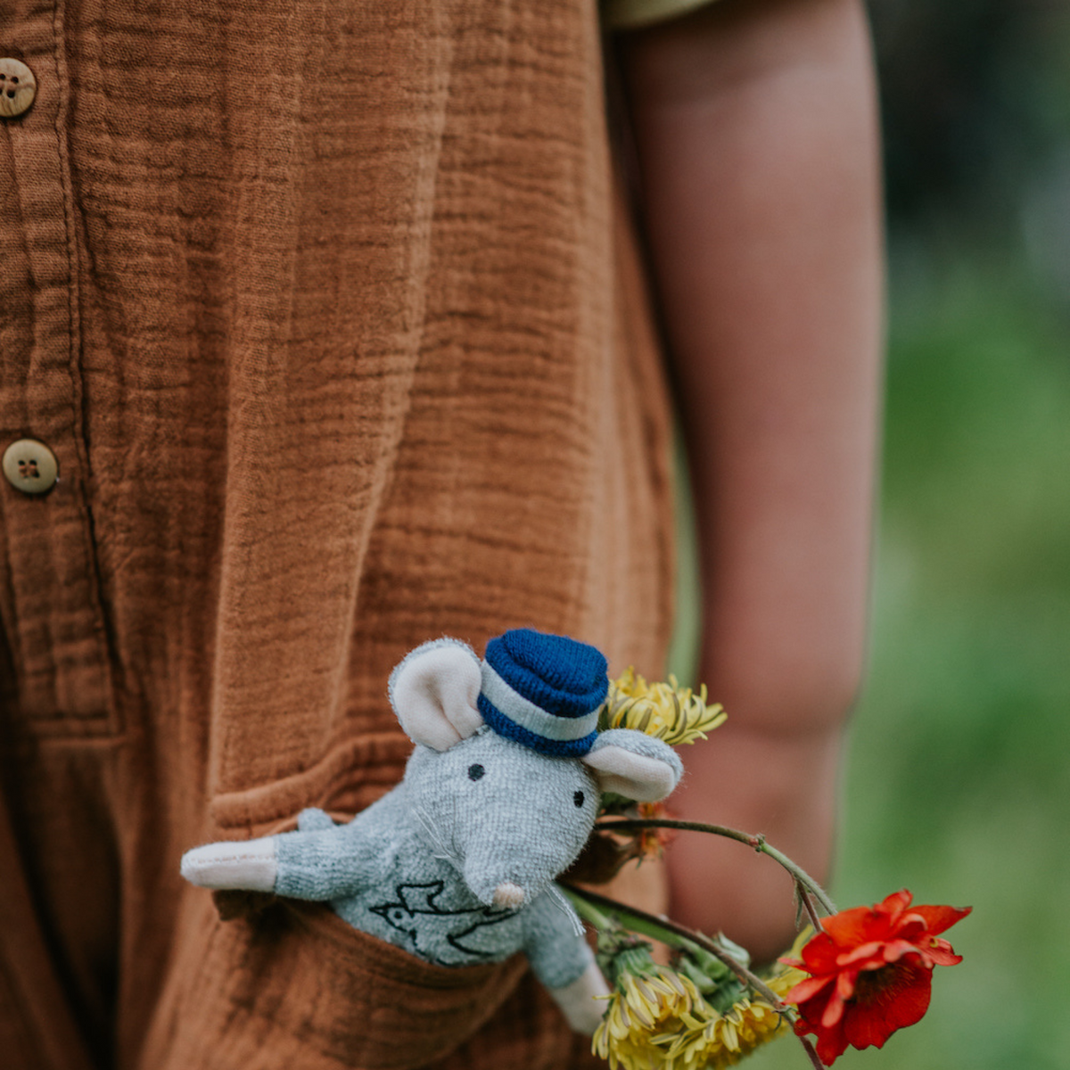 Small plush mouse toy with a blue hat held by a person wearing a brown shirt outdoors.