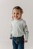 A young girl with brown hair in a plaid hair tie, wearing a pale sage green long-sleeve top with floral embroidery on the sleeves and blue jeans, standing against a light gray studio background.