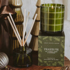 A green glass candle on top of its branded box next to a reed diffuser bottle with wooden reeds. The candle is in a translucent olive-green glass with a white windowpane plaid pattern, and the box reads 'FRASER FIR' by Sweet Water Scents.
