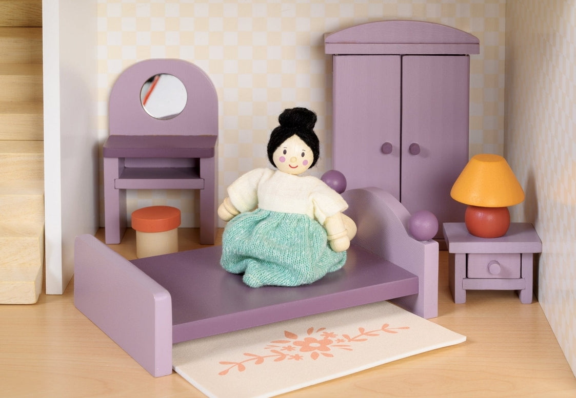 A dollhouse bedroom scene featuring a wooden doll sitting on a double bed with a purple frame. Next to the bed is a bedside table with a yellow-shaded lamp, a wardrobe against the wall, a dressing table with a mirror and stool, and a small floral rug on the floor.