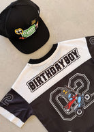 A black and white mesh baseball-style jersey and matching black cap laid flat on a light concrete surface. The cap has a colorful 'Happy Birthday' graphic, and the jersey features 'BIRTHDAYBOY' across the back with a large number 2 and a cartoon racer graphic.