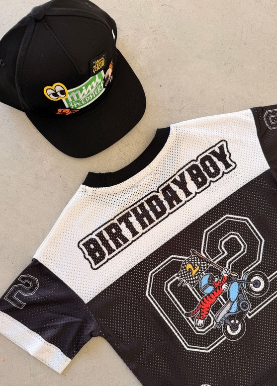 A black and white mesh baseball-style jersey and matching black cap laid flat on a light concrete surface. The cap has a colorful 'Happy Birthday' graphic, and the jersey features 'BIRTHDAYBOY' across the back with a large number 2 and a cartoon racer graphic.