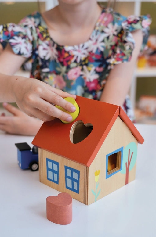 A young child looks down intently as they play with a wooden shape sorter house on a table, with an adult or older child leaning in beside them to watch. Colorful wooden blocks are scattered on the surface near the toy.