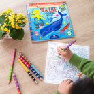 Child coloring a sea life coloring book with colored pencils on a wooden table.