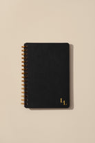 Black notebook with gold spiral binding and initials on a beige background