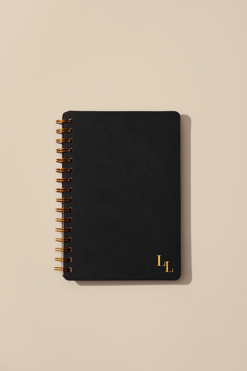 Black notebook with gold spiral binding and initials on a beige background