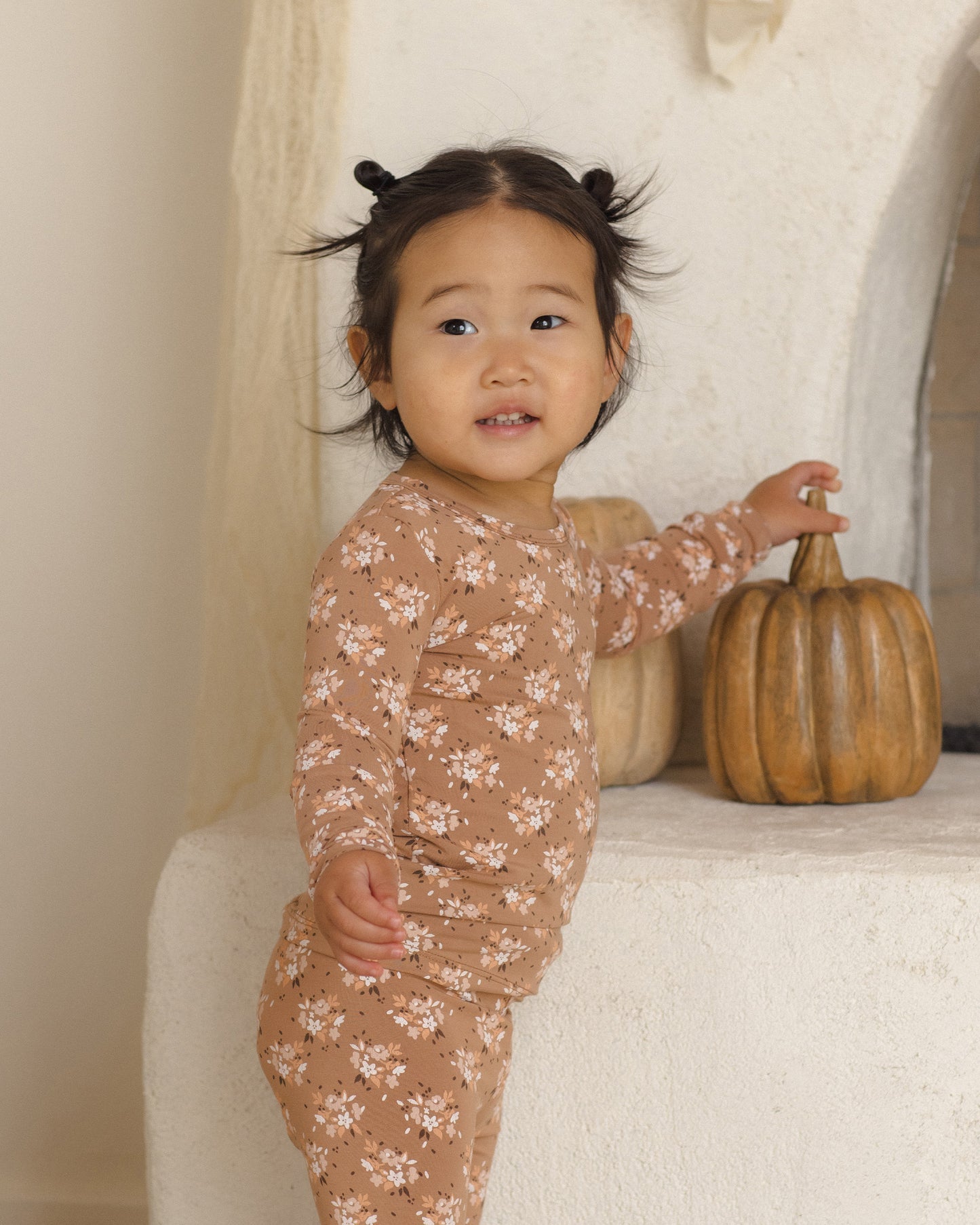 A toddler girl wearing a long-sleeve pajama set in a warm tan color with a delicate white floral pattern, standing next to a small decorative pumpkin on a textured white stone shelf.