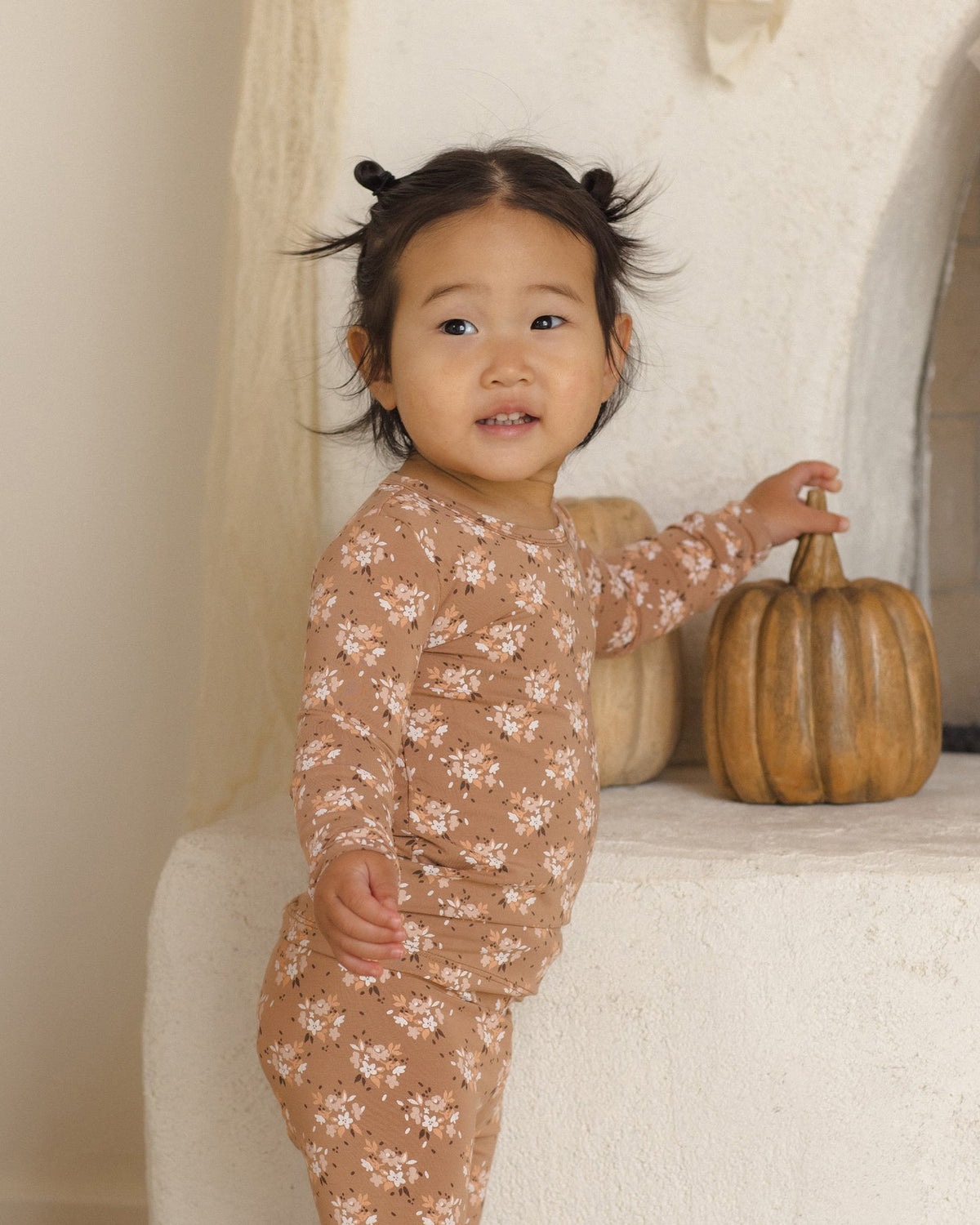 A toddler girl wearing a long-sleeve pajama set in a warm tan color with a delicate white floral pattern, standing next to a small decorative pumpkin on a textured white stone shelf.
