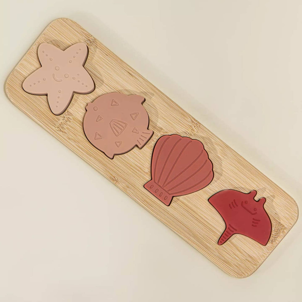A wooden toddler puzzle board featuring four sea-themed shapes: a starfish, a fish, a clam shell, and a conch shell in muted pink, peach, dusty rose, and red tones, displayed on a light cream background.