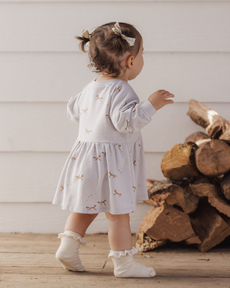 A toddler wearing the Rylee + Cru Foxes Raglan Dress in a soft fog hue, styled with a sweet matching bow and ruffled socks.