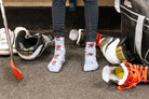A person wearing light blue crew socks with red and white cartoon graphics stands on asphalt surrounded by sports gear, including shoes, a helmet, gloves, and a bag.