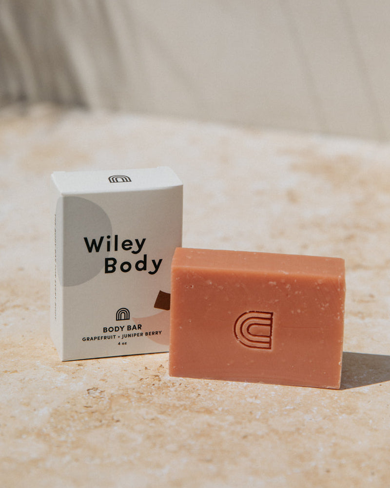 A rectangular Wiley Body body bar in a warm peachy pink color stands in front of its white packaging box on a light beige stone surface.