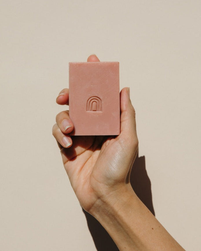 A hand holding a rectangular dusty pink soap bar with an embossed arch logo, set against a solid beige background.