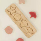 A wooden four-piece puzzle board with engraved star, circle, shell, and fish shapes, surrounded by matching pink and red plastic puzzle pieces on a light cream background.