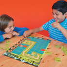 Two children playing a board game on a wooden table with an orange wall in the background.
