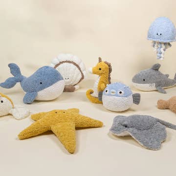 A collection of soft, crocheted sea creature plush toys including a blue whale, yellow starfish, gray shark, blue jellyfish, and other ocean-themed stuffed animals arranged on a light beige background.