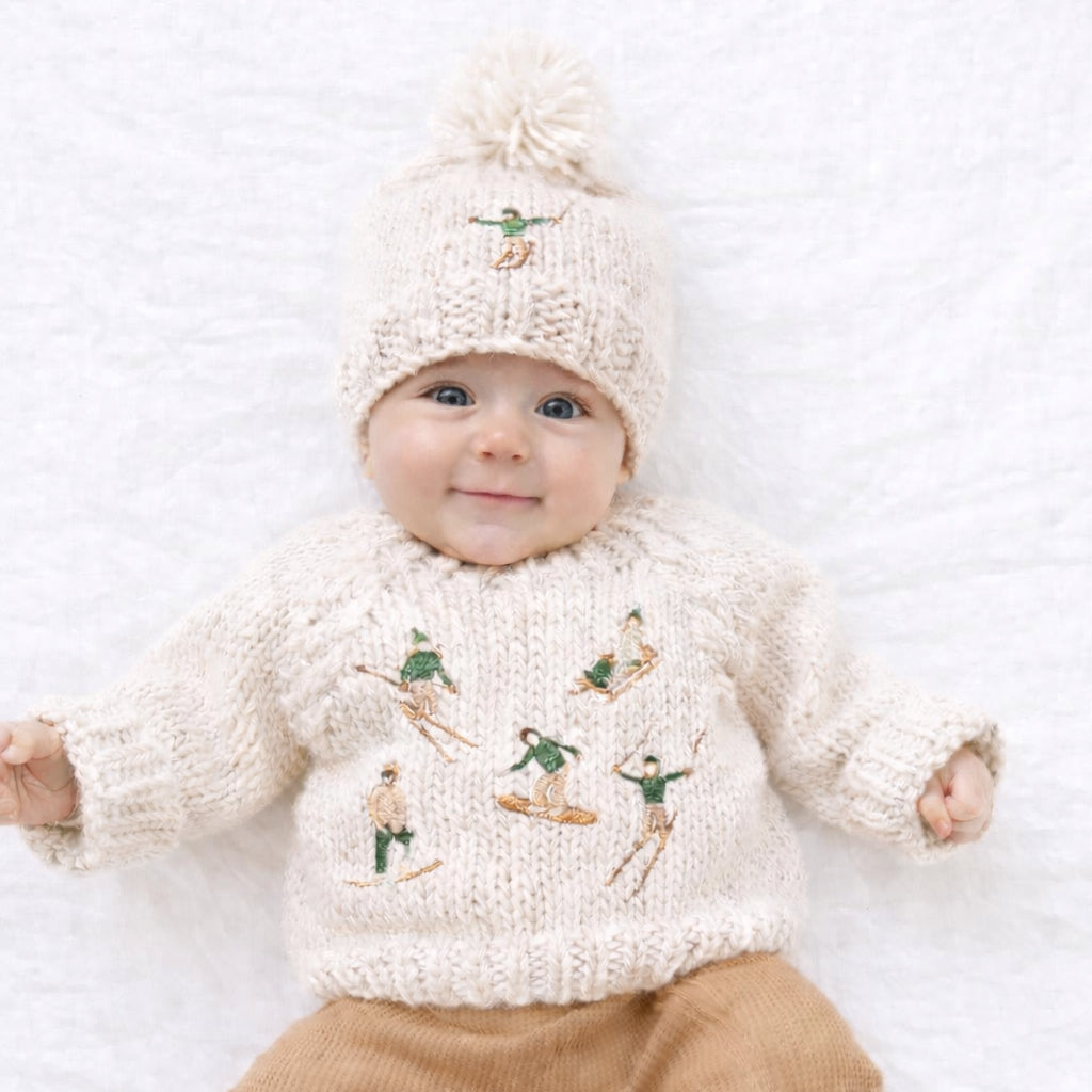 Baby wearing a cream sweater with embroidered designs and a matching hat on a white background