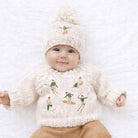 Baby wearing a cream sweater with embroidered designs and a matching hat on a white background