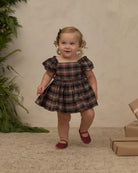 A toddler girl wearing a brown and black plaid dress with puff sleeves and maroon shoes, standing in a studio setting with a textured beige backdrop, greenery on the left, and wrapped gifts on the right.