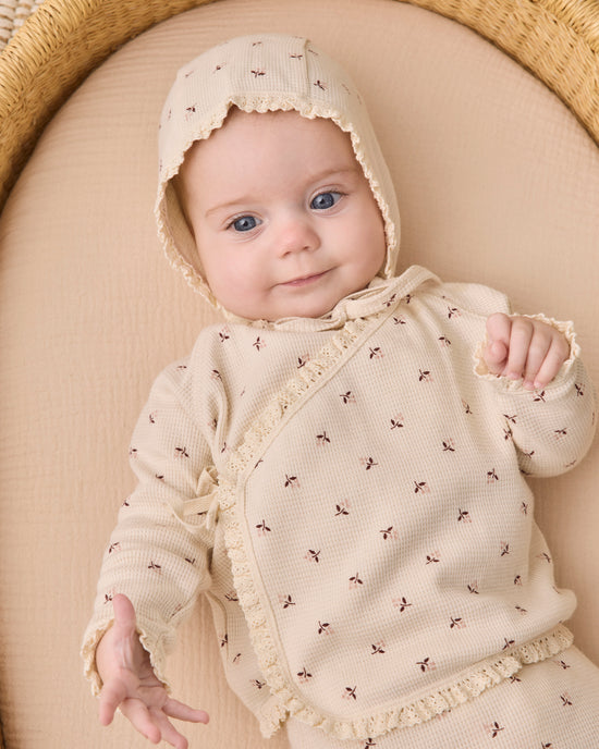 A baby wearing a cream-colored knit romper and matching bonnet with small brown embroidered motifs, lying on a beige cushion inside a woven basket.