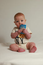 A baby sitting on a white surface against a light beige background, wearing a cream-colored short-sleeve bodysuit with black lettering and holding a blue cereal box.