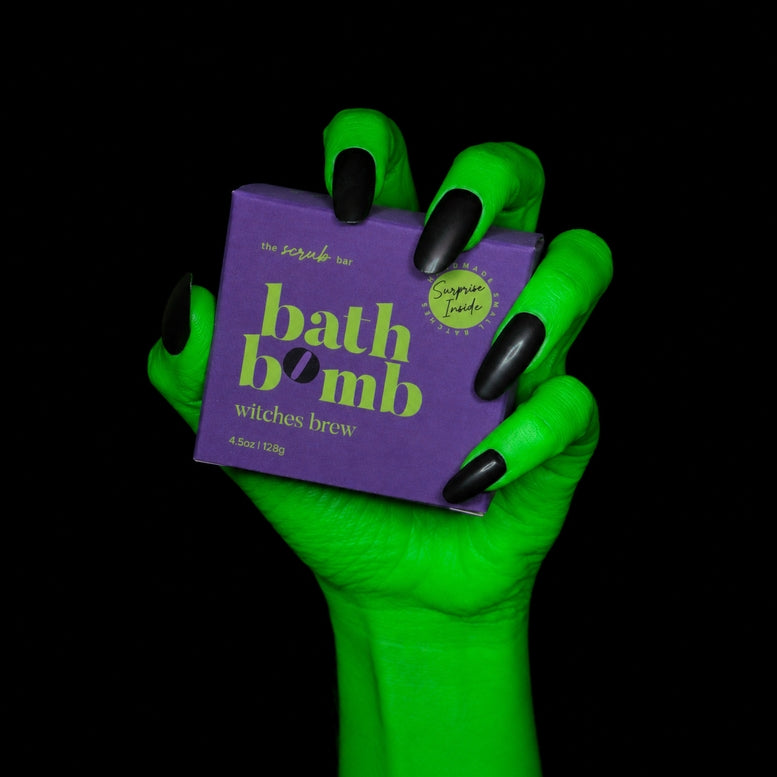 A hand with bright neon green skin and black fingernails holds a purple box of 'bath bomb witches brew' by The Scrubba Bar against a solid black background.