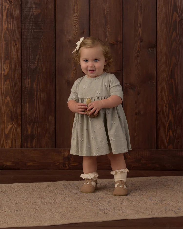 A toddler wearing the sage-hued Rylee + Cru Finn Dress in their charming mushrooms print, styled with a matching hair bow and ruffled socks.