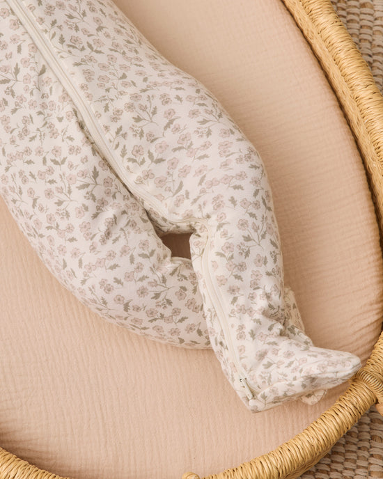 A close-up of a footed baby sleeper with a delicate beige and white floral pattern, resting on a peach-colored muslin mattress inside a woven rattan bassinet.