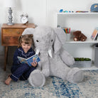 Child sitting on a large gray stuffed elephant toy on a carpeted floor.