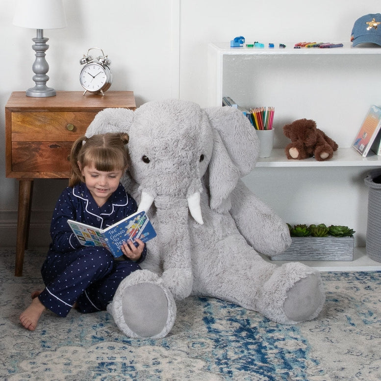 Child sitting on a large gray stuffed elephant toy on a carpeted floor.