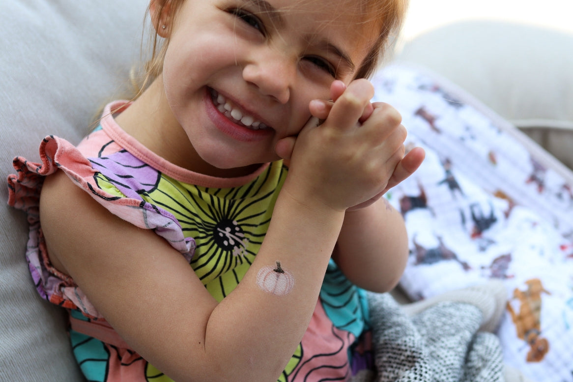 A young girl smiling while wearing a small temporary pumpkin tattoo on her inner forearm, lying on a couch with a patterned blanket in the background.