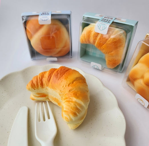 A styled product display of various bakery items, including golden-brown croissants, round buns, and braided bread, with some packaged in boxes and others arranged on a beige fabric surface.