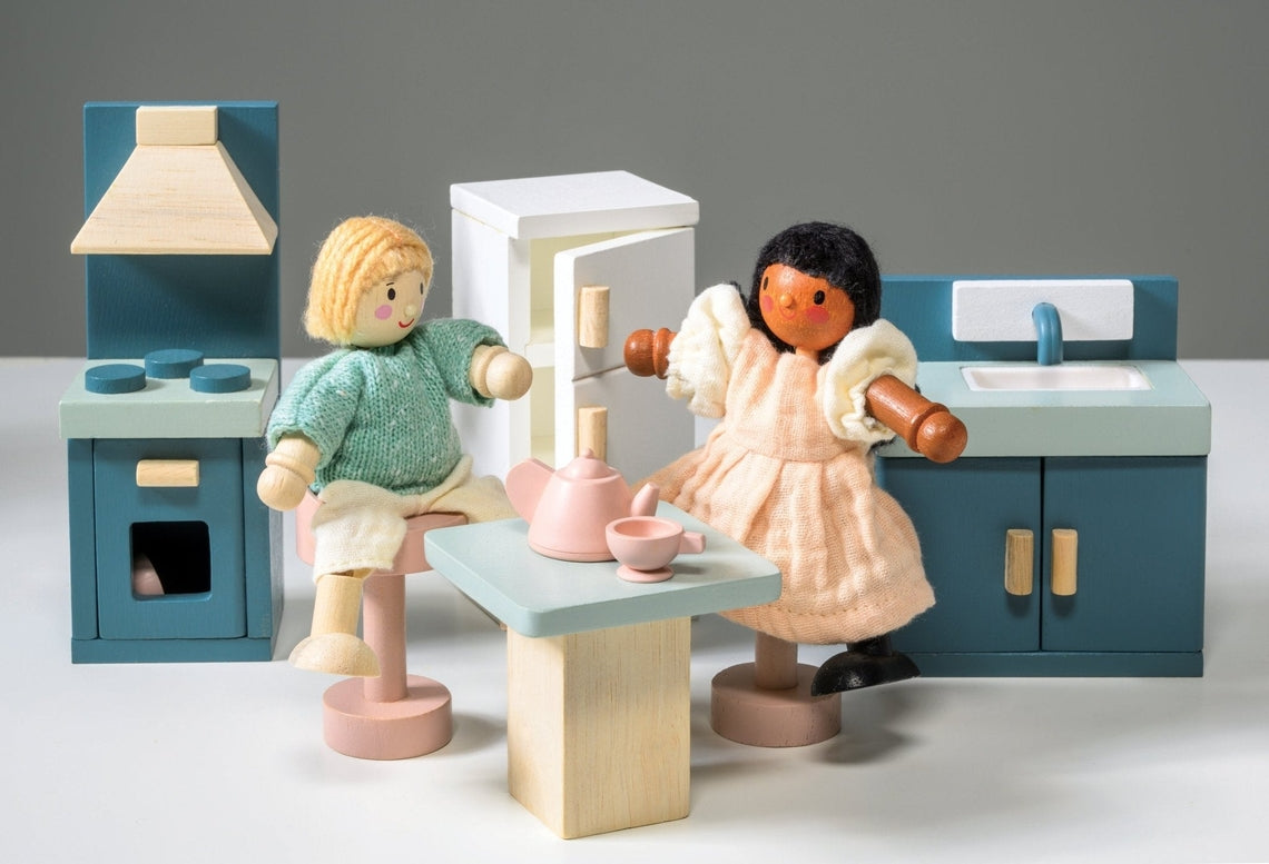 Wooden kitchen furniture set by Mentari against a white background, featuring two wooden dolls interacting at a light wood-colored table set with a teapot and two cups, surrounded by a blue stove with an oven and hood, a white refrigerator with wooden handles, and a blue sink unit with a cupboard.