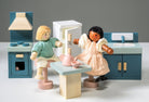 Wooden kitchen furniture set by Mentari against a white background, featuring two wooden dolls interacting at a light wood-colored table set with a teapot and two cups, surrounded by a blue stove with an oven and hood, a white refrigerator with wooden handles, and a blue sink unit with a cupboard.
