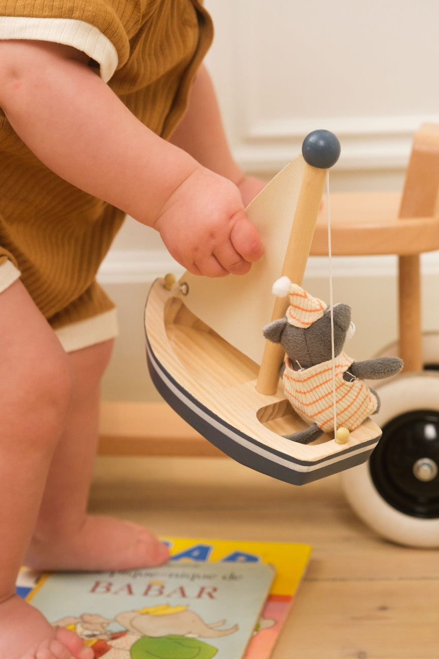 Child holding a wooden toy boat with a plush inside, on a wooden floor.