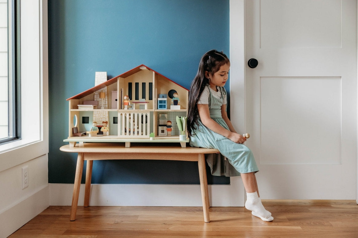 A young child sitting on a white bench by a teal wall plays with the Wooden Cedar Chalet Modernist Dollhouse by Mentari, which is arranged with miniature furniture and figures inside its open-front mid-century modern home.