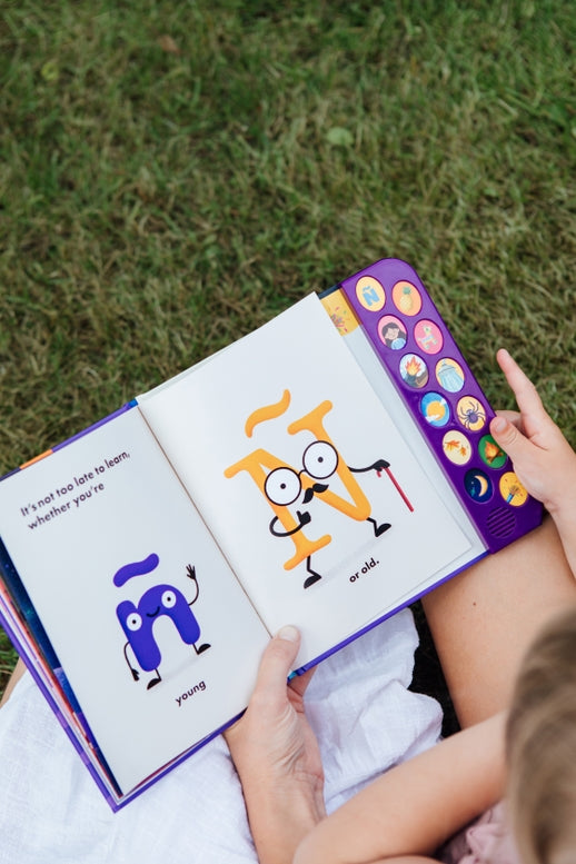 A child holding open "What Does Ñ Sound Like?: A Spanish-English Sound Book," showing an interior illustrated page featuring two animated letter characters and the text "It's not too late to learn whether you're young... or old," with a vertical panel of sound buttons on the right edge.