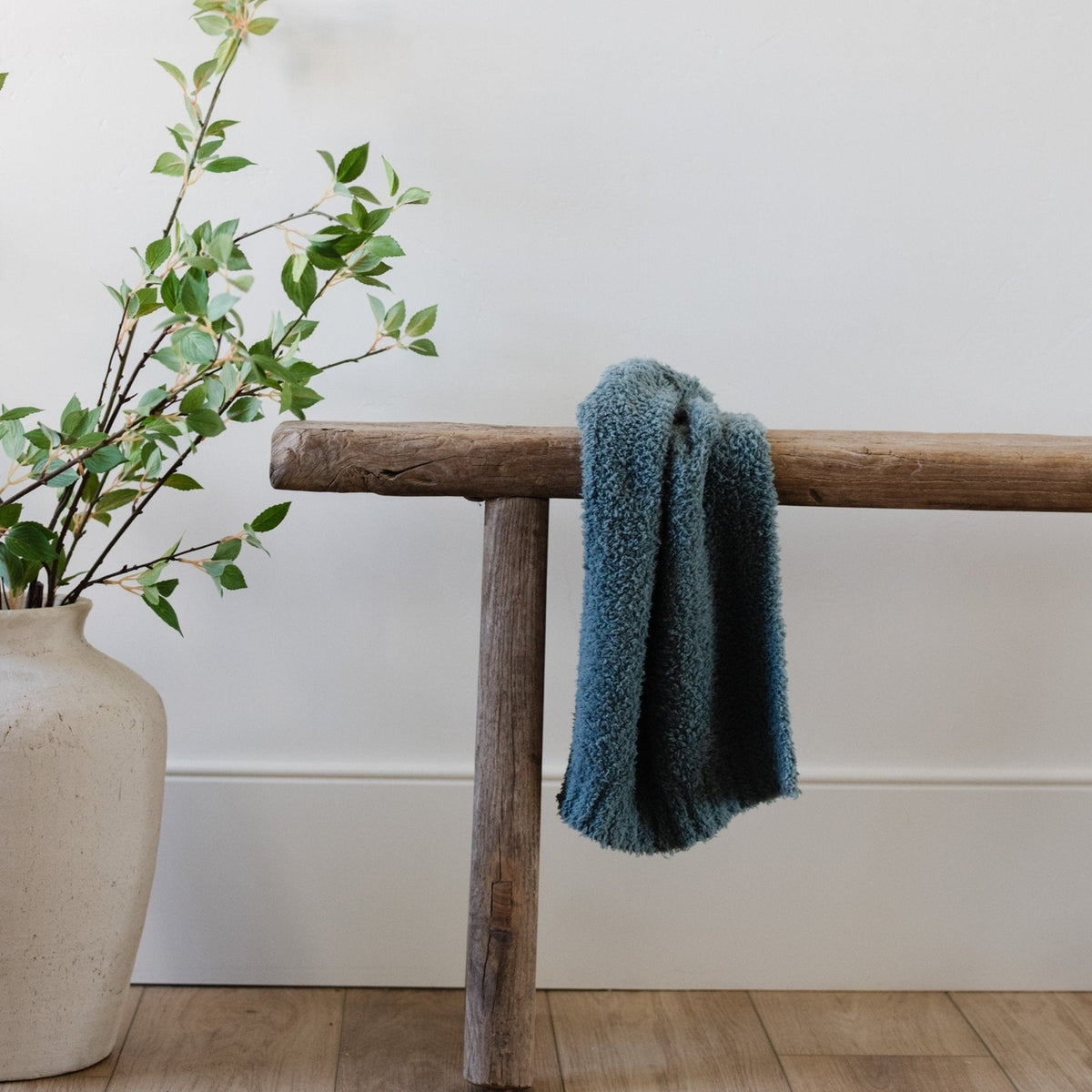 Blue textured towel draped over a wooden bench with a vase of greenery on a white wall background