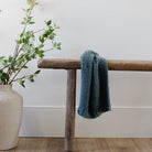 Blue textured towel draped over a wooden bench with a vase of greenery on a white wall background