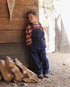 A young boy wearing blue denim overalls and a red-and-white striped long-sleeve shirt leans against a rustic wooden wall next to a pile of firewood in an outdoor setting.