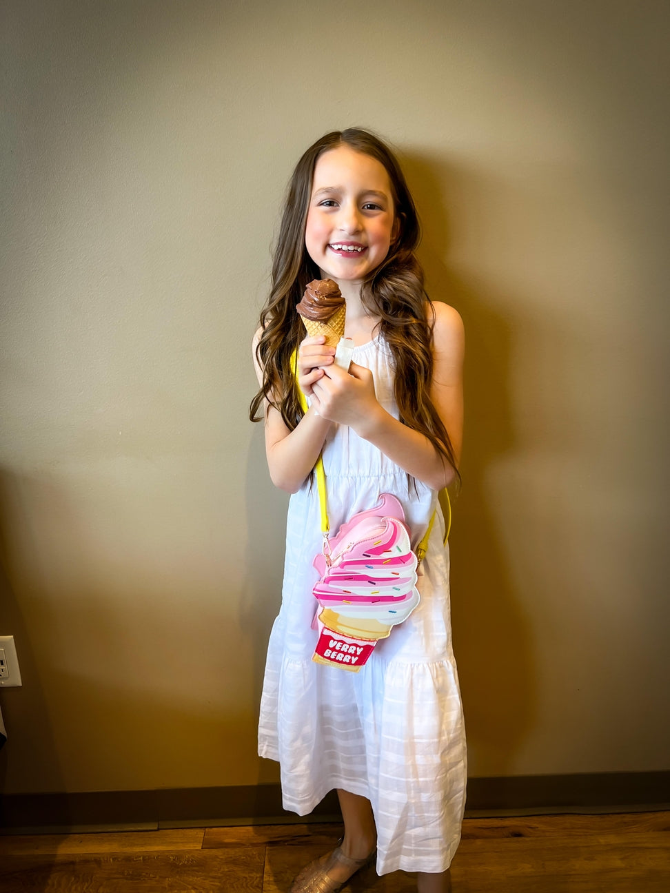 Young girl in a white dress holding a toy ice cream cone against a plain wall.