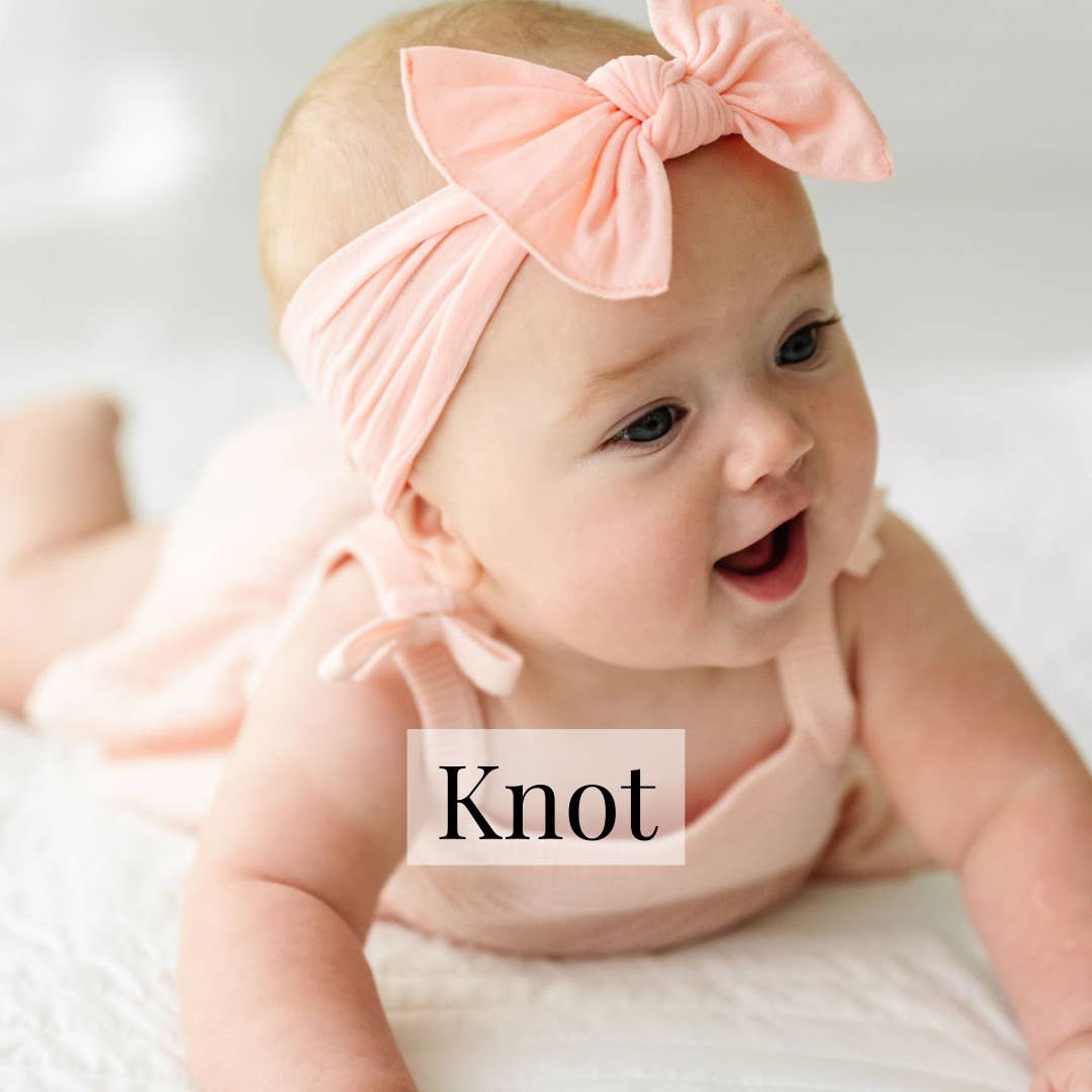 A baby lying on a white blanket wearing a pale pink knit bow headband and matching pink outfit. The word 'Knot' appears in a white box over the center.