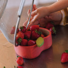 Child's hand reaching into a pink bowl filled with strawberries on a wooden surface.