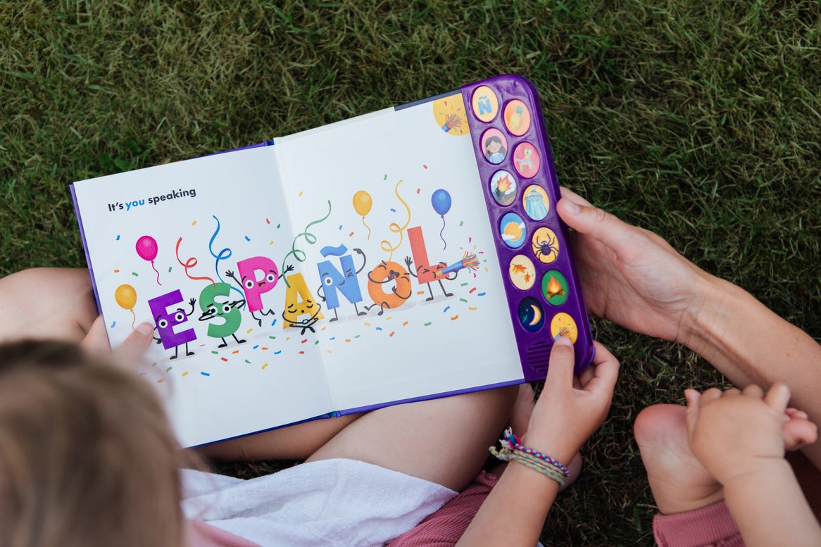 A child and adult holding open "What Does Ñ Sound Like?: A Spanish-English Sound Book," showing an interior colorful spread with animated letters spelling "ESPAÑOL," balloons, and a vertical panel of interactive sound buttons on the right edge.