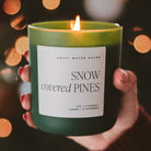 Hand holding a 'Snow Covered Pines' candle with a blurred light background