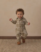 A toddler with curly brown hair wearing a beige and brown plaid button-up romper and brown leather shoes, walking forward on a patterned rug against a beige studio backdrop.