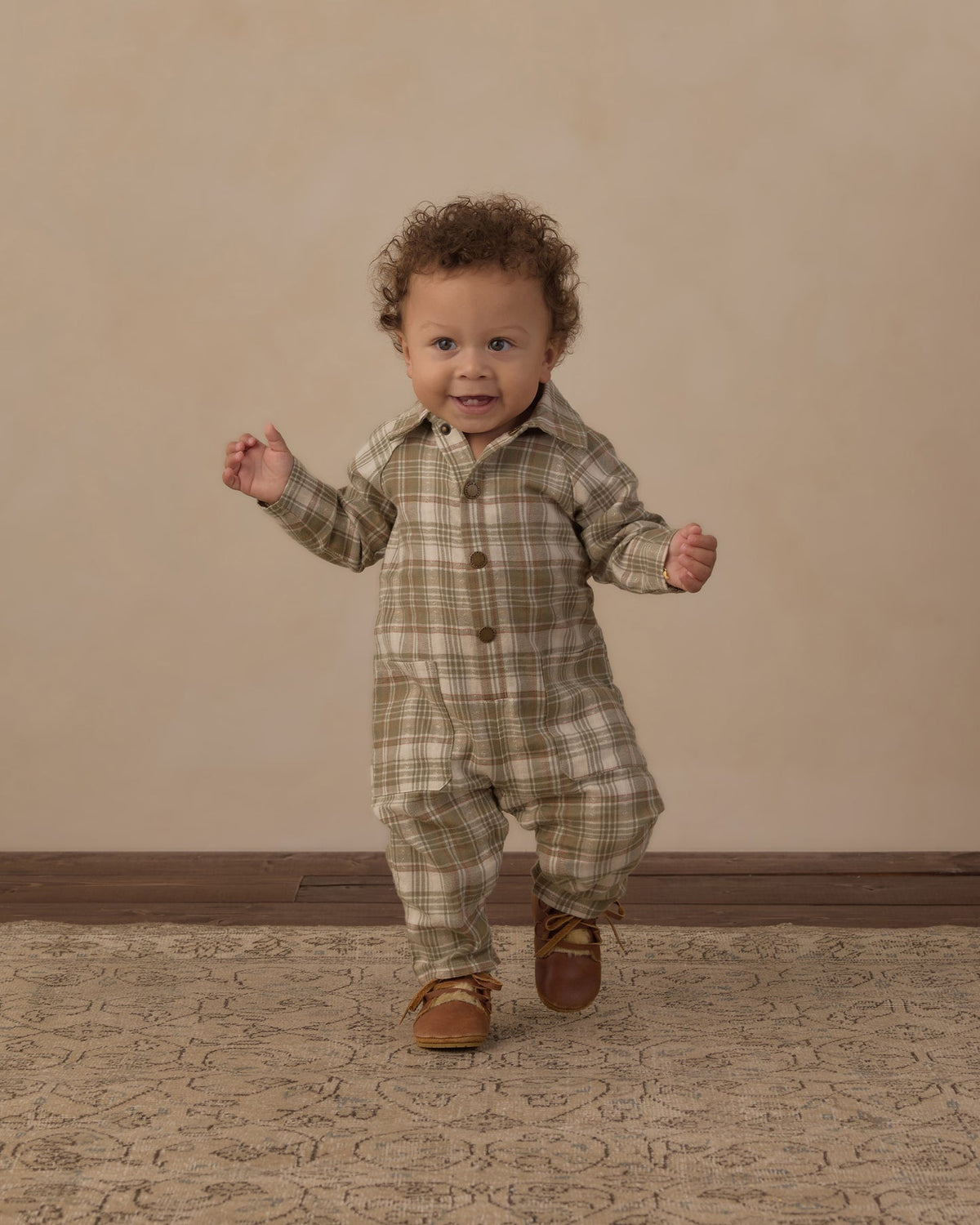 A toddler with curly brown hair wearing a beige and brown plaid button-up romper and brown leather shoes, walking forward on a patterned rug against a beige studio backdrop.